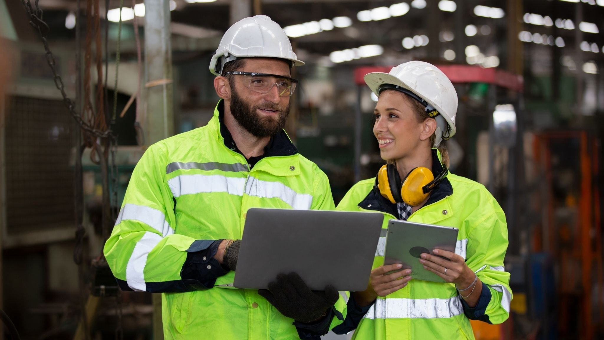Operations man an women smiling, with laptop, hi vis and hard hats. Factory setting